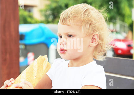 Outdoor closeup portrait of cute Caucasian blond baby girl eating big French baguette Stock Photo