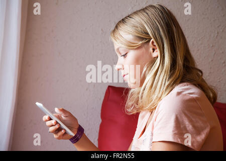 Cute Caucasian blond teenage girl in pink t-shirt using smartphone for messaging, indoor closeup profile portrait Stock Photo