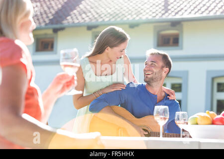 Smiling young couple playing guitar and drinking wine on sunny patio Stock Photo