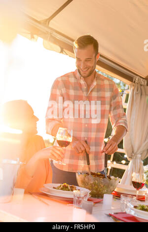 Young man serving salad to wife drinking wine at sunny patio table Stock Photo