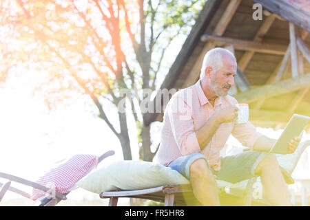Senior man drinking coffee and using digital tablet on lounge chair in backyard Stock Photo