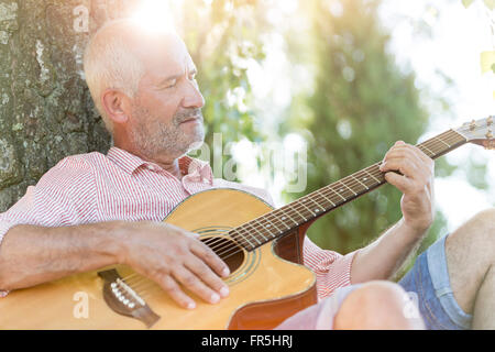 Senior man playing guitar against tree trunk Stock Photo