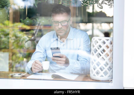 Businessman drinking espresso and checking cell phone at cafe window Stock Photo