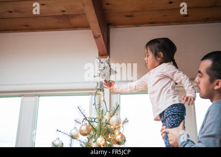 Father lifting toddler girl putting star on Christmas tree Stock Photo