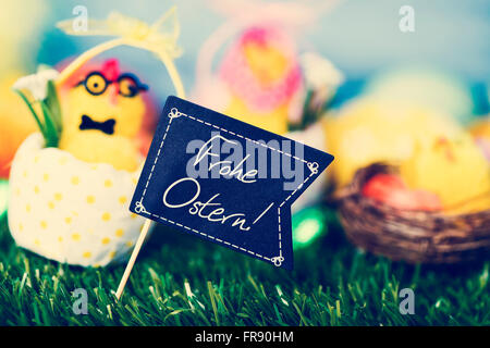 a funny teddy chick, emerging from a dot-patterned eggshell, with a black flag-shaped signboard with the text text Frohe Ostern, Stock Photo