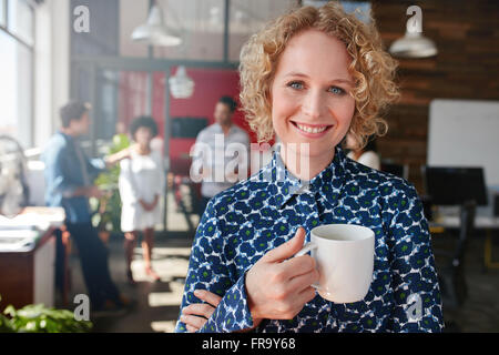 Portrait of happy young businesswoman holding a cup of coffee looking at camera and smiling. She is standing in her office with Stock Photo