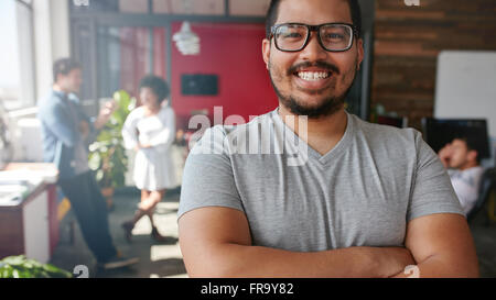 Smiling male designer looking at camera at office. Mixed race male creative professional with colleagues talking in background. Stock Photo