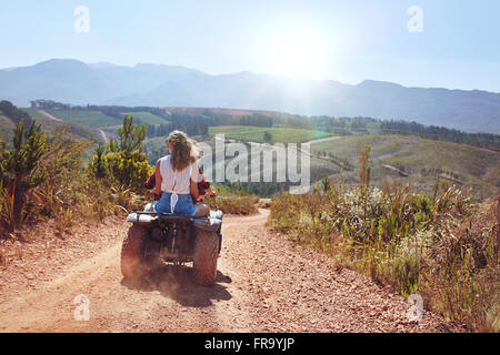 Rear view shot of young woman sitting behind her boyfriend driving a quad bike on a sunny day. Couple in countryside riding on a Stock Photo