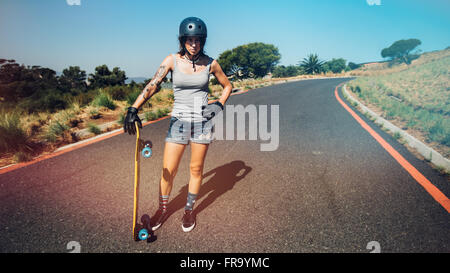Full length portrait of young woman standing on a countryside highway with a longboard. Stock Photo