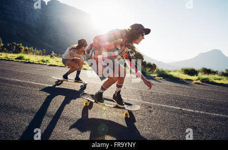 Side portrait of young people skateboarding together on road. Young man and woman longboarding down the road on a sunny day. Stock Photo