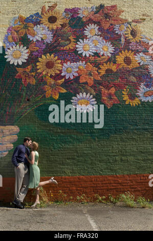 A couple kisses as they lean against a wall with a large mural of a colourful bouquet of flowers painted on the wall Stock Photo