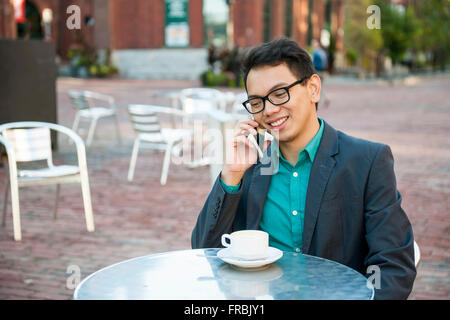 Successful young asian man in business casual attire sitting and smiling in relaxing outdoor cafe with cup of coffee talking on Stock Photo