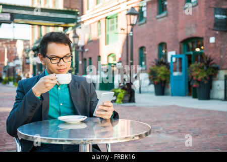 Serious young asian man in business casual attire sitting in outdoor cafe drinking cup of coffee while using mobile phone, with Stock Photo
