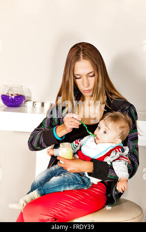 Young beautiful mother is feeding her cute  little boy with a puree in modern surrounding. Stock Photo