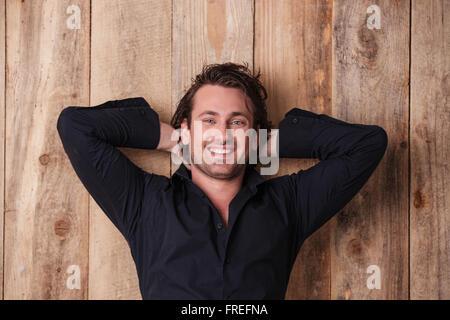 Happy relaxed young man in black shirt posing with hands behind head over wooden background Stock Photo