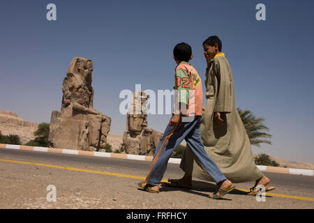 Young Egyptian boys walks past the ancient Egyptian Colossi of Memnon site, Luxor, Nile Valley, Egypt. The Colossi of Memnon (memorial temple of Amenophis III) are two massive stone statues of Pharaoh Amenhotep III, who reigned during Dynasty XVIII. For the past 3,400 years (since 1350 BC) they have stood in the Theban necropolis, west of the River Nile from the modern city of Luxor. Stock Photo