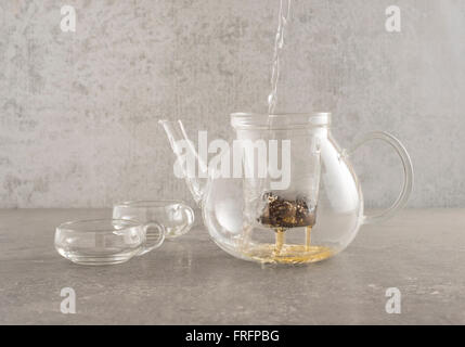 Hot water pouring in glass teapot and sifting through tea leaves. Two glass cups are placed on the side on a stone kitchen table Stock Photo