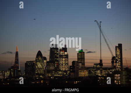 The sun is setting on the City of London and the lights are on, seen from Hackney, East London. A helicopter hovers above.