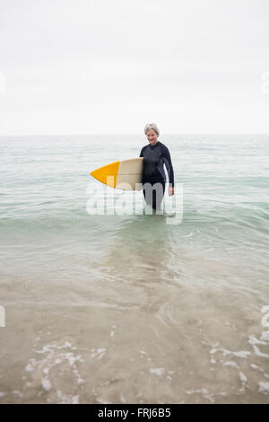 Senior woman in wetsuit standing in water with surfboard on the beach Stock Photo