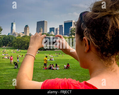Woman using a smartphone to take a photo of people relaxing on the grass in Central Park, New York City, with the Manhattan skyline in the background. Stock Photo