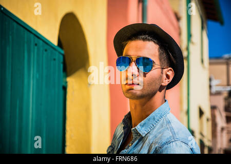 Portrait of dark haired young man in blue mirrored sunglasses and black hat. Cityscape on background, with colorful houses in It Stock Photo