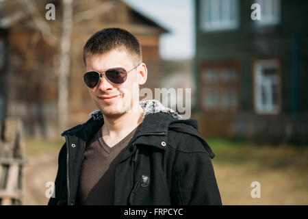 Young Handsome Man Staying Near Old Wooden House In Sunny Spring Day. Casual Style - Jacket, Sunglasses. Stock Photo