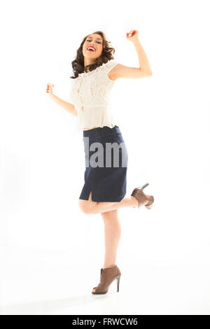 Beautiful young woman on one leg, in white blouse and blue denim skirt, full length in brown heels, arms raised, head back. Stock Photo