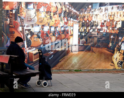 Old man sat on a public bench in front of a boarded up shop, covered with a photo of the interior of a music shop, Havant, Hants Stock Photo