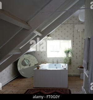 Circular mirror on floor beside bath set in the center of a nineties attic bathroom with patterned wallpaper Stock Photo