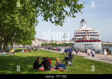 People enjoying a sunny Sunday afternoon in the park with cruise ship MS Deutschland Reederei Peter Deilmann moored along La Garonne river, Bordeaux, Gironde, Aquitane, France Stock Photo