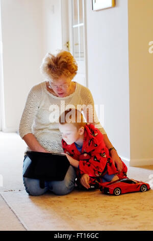 A grandmother with her grandchild both sharing an iPad or tablet whilst cuddled together. They’re illuminated by the screen on the device Stock Photo