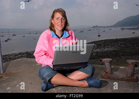 A woman in a pink fleece sitting crossed-legged on the beach at Beaumaris,Anglesey,Wales,UK,using her laptop computer. Stock Photo