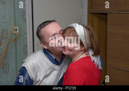 Senior man in white knitted vest is kissing on the cheek his smiling girlfriend in bright red blouse. Stock Photo