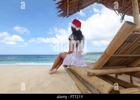 Woman in Santa Claus hat sitting at chaise lounge with straw parasol on white sandy beach at Philippines Stock Photo