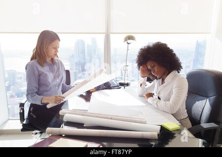 Team of two female architects, sitting at desk in office skyscraper. The women talk reviewing a building plan. Medium shot Stock Photo