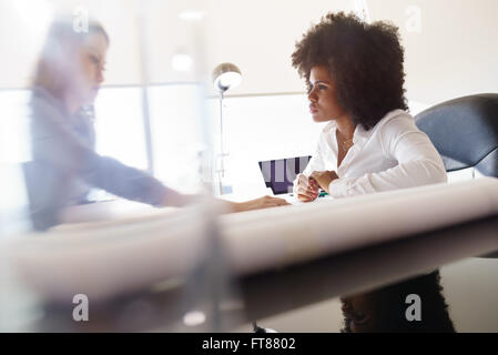 Team of two female architects, sitting at desk in office. The women talk reviewing a building plan. Medium shot Stock Photo