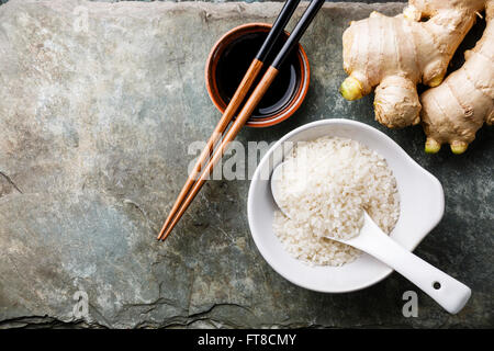 Raw white rice, fresh ginger root, soy sauce and sushi chopsticks on gray stone slate background Stock Photo