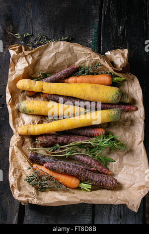 Heap of colorful raw carrots with fresh thyme herbs on paper bag over black wooden surface. Top view.