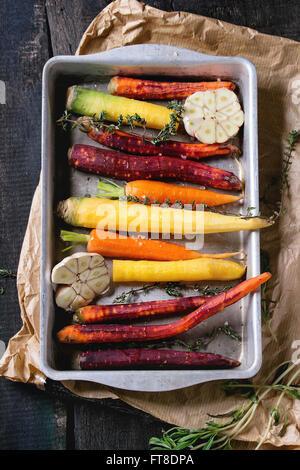 Ready for baking peeled colorful raw carrots with fresh thyme herbs and sliced garlic in aluminum tray on paper bag over black w