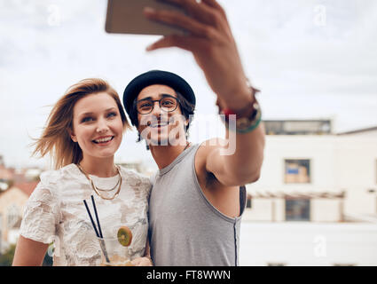 Two young friends taking self portrait with smart phone during a party. Young man and woman taking selfie at rooftop party. Stock Photo