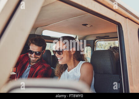Young couple sitting in their car during roadtrip. Young man and woman wearing sunglasses sitting inside the car and smiling. Stock Photo