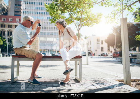 Side view of senior couple taking pictures while on vacation. Mature couple sitting outdoors on a bench photographing each other Stock Photo