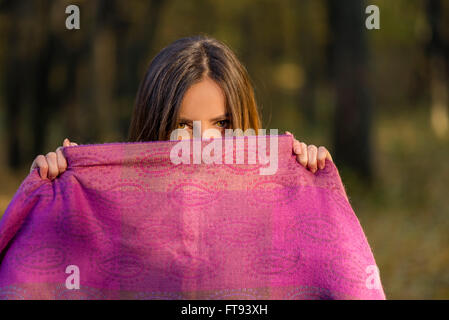 Behind purple scarf. Pretty teenager girl with brown eyes is covering her face with the purple scarf in the afternoon woods. Stock Photo