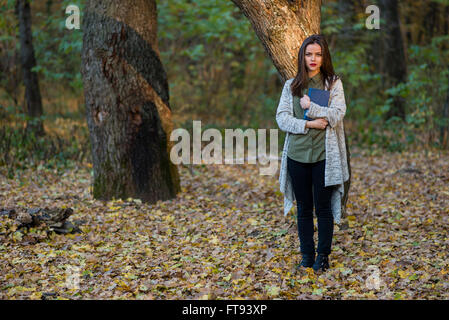 Scared by reading fairytales. A teenager girl is holding a book in an evening forest with two oak trees in the background. Stock Photo