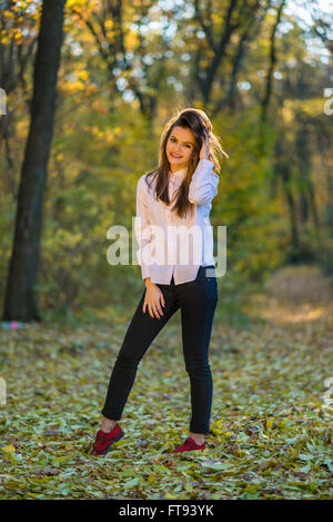 A smiling good looking girls is posing in a pink shirt, dark blue jeans and red shoes in an autumn forest. Stock Photo