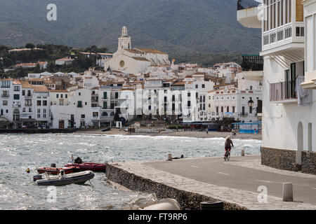 Harbour and Village of Cadaques. A woman bikes along the harbour road in Cadaques.  The town's church rises above the village Stock Photo