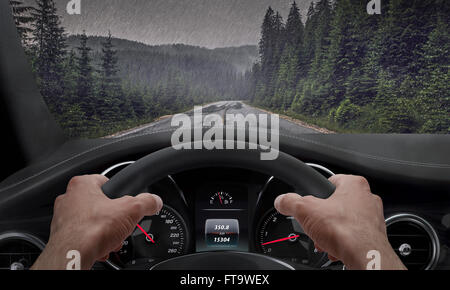 Driving in rainy weather. View from the driver angle while hands on the wheel. Rain splashed windshield. Stock Photo