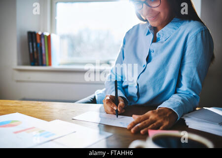 Selective focus close up of cheerful young self-employed woman wearing glasses and blue shirt at desk writing on form next to cu Stock Photo