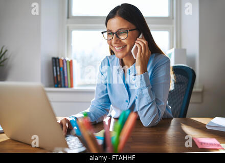 Single happy female business owner with smile and eyeglasses on phone and working on laptop computer at desk with bright window Stock Photo