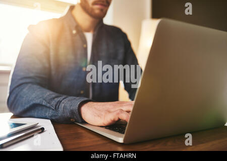 Businessman typing in data on the laptop computer as he sits at his desk in the office backlit by bright sun flare, close up on Stock Photo